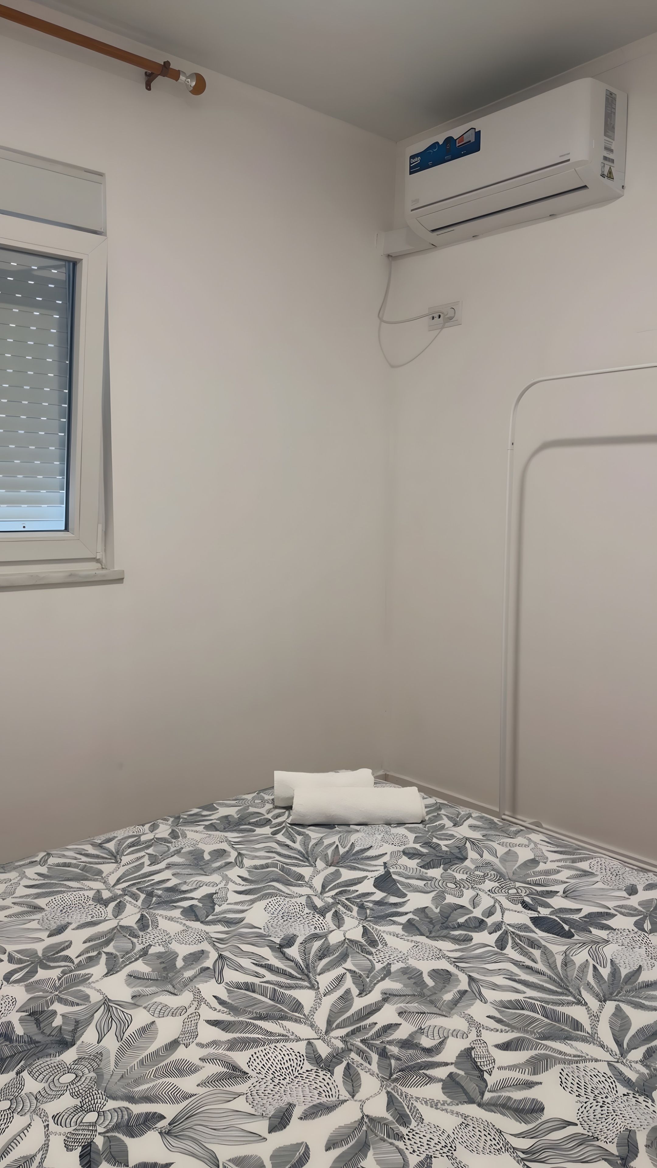 A room with a bed, an air conditioning unit, a window with blinds, and a wall-mounted radiator.