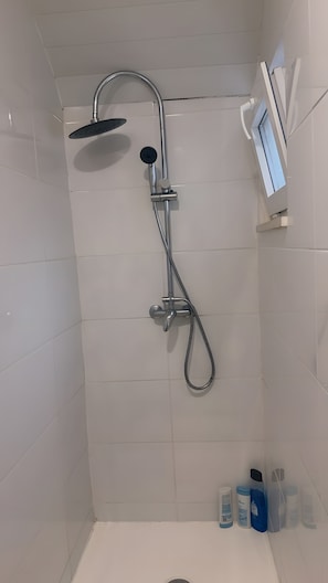 En bruser med håndbruser og vandhane på væggen, et lille vindue og toiletartikler på en hylde.