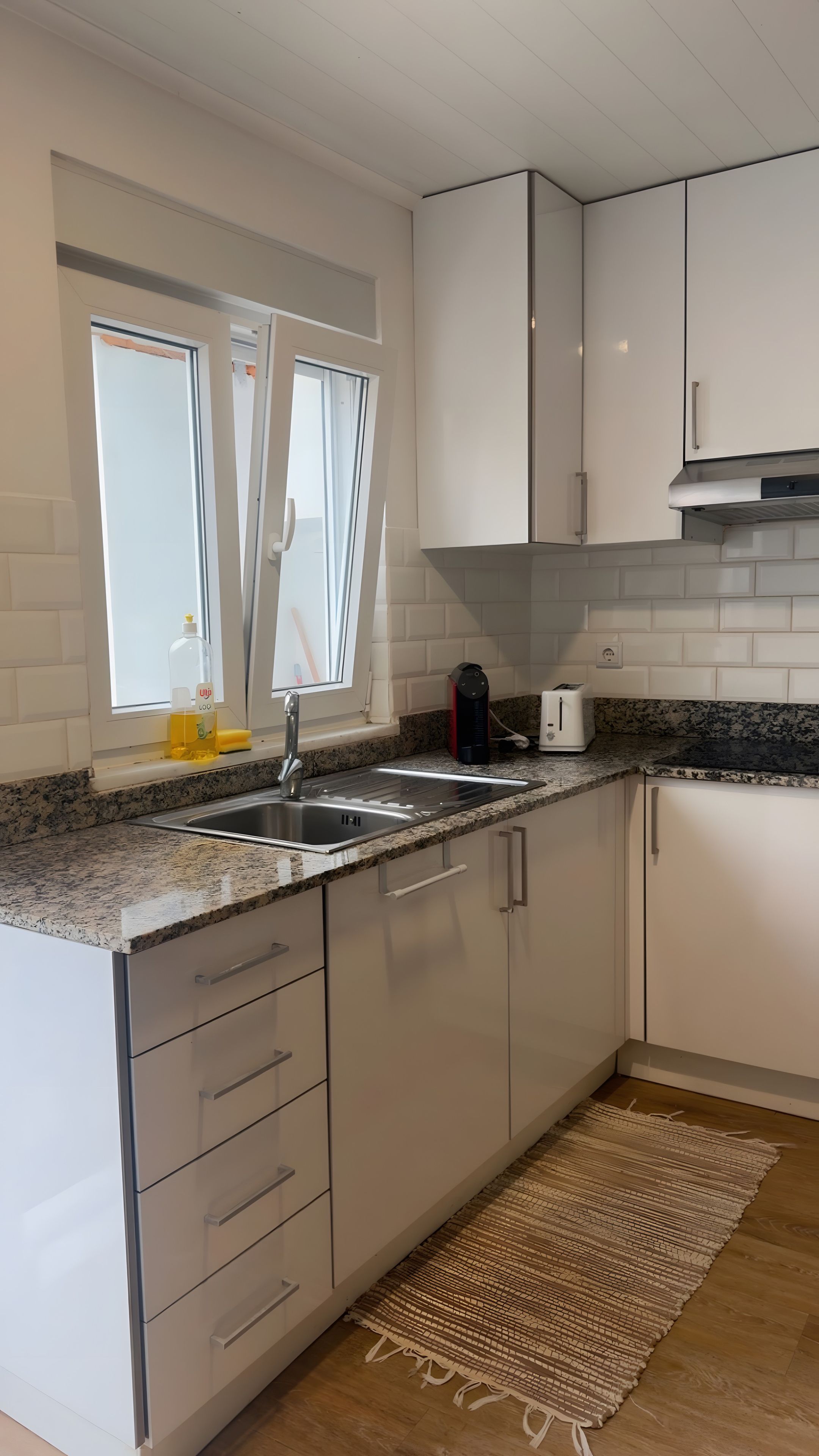 Una cocina moderna con armarios blancos, encimera de granito, pileta de acero inoxidable y una ventana con cortinas.