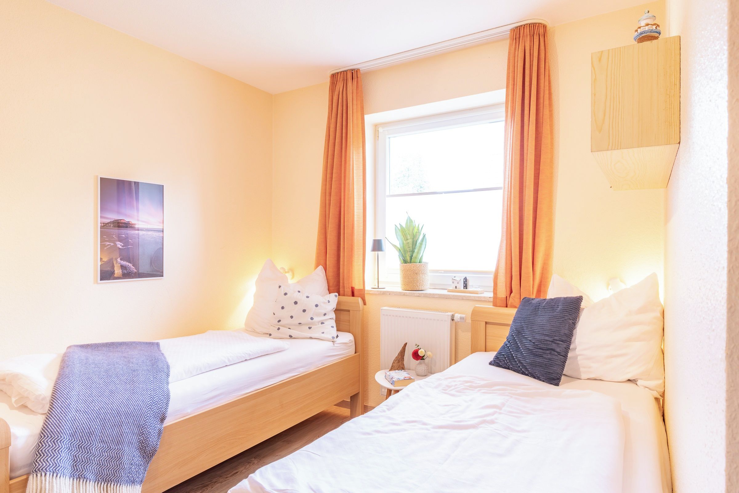 Een hotelkamer met twee eenpersoonsbedden, een raam met gordijnen, een radiator, een klein tafeltje met een plant en een wandlamp.
