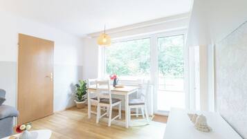Una habitación luminosa con suelo de madera, una mesa blanca con sillas, una lámpara colgante y vistas al exterior con vegetación.