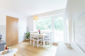 Una habitación luminosa con suelo de madera, una mesa blanca con sillas, una lámpara colgante y vistas al exterior con vegetación.