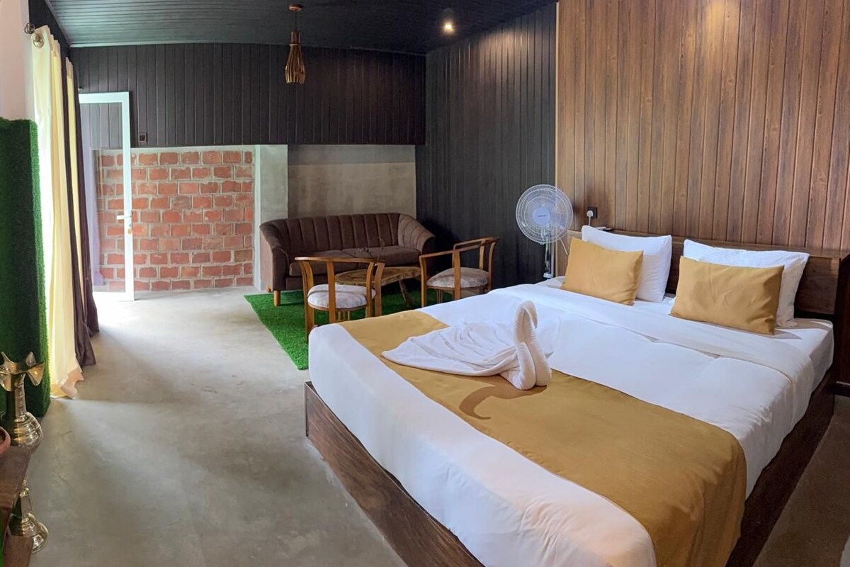 A hotel room with a large bed, a sitting area with a table and chairs, a fan, and a brick wall in the background.