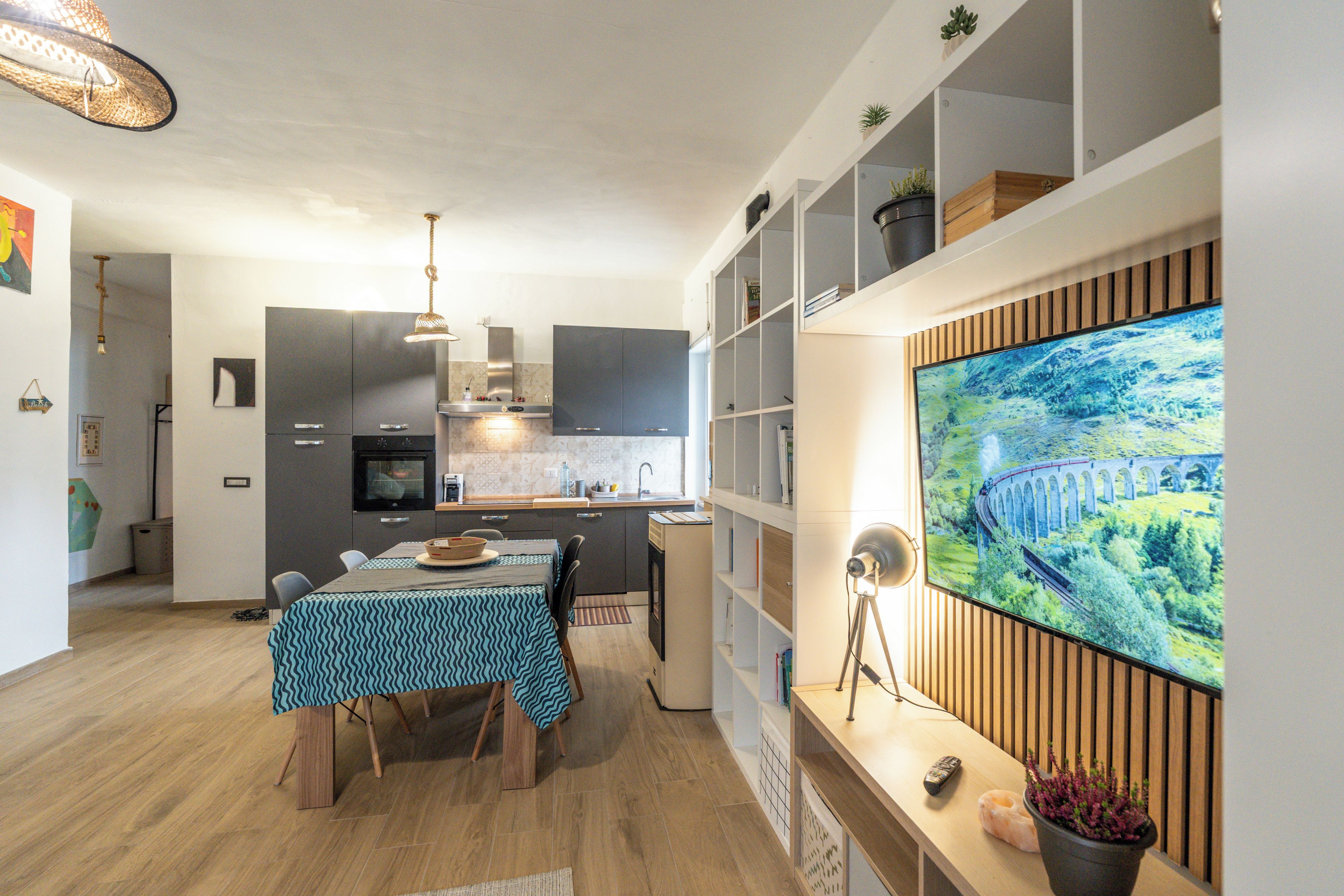 A modern kitchen with a dining area, built-in shelving, and a large flat-screen TV mounted on the wall.