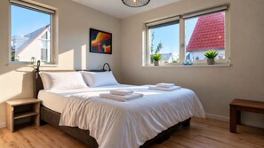 A bedroom with a bed, bedside tables, a window with a view of buildings, and a painting on the wall.