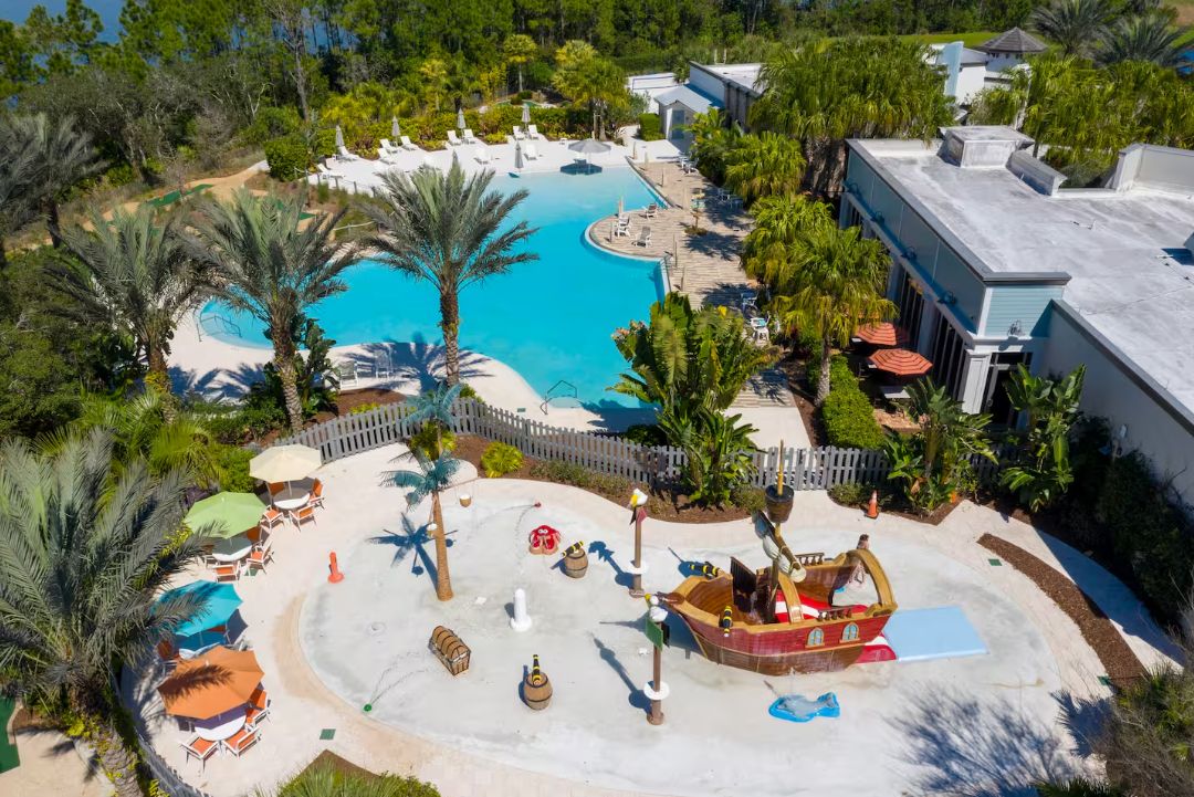 An aerial view of a resort with a large pool, palm trees, and a beach area with sun loungers and umbrellas.