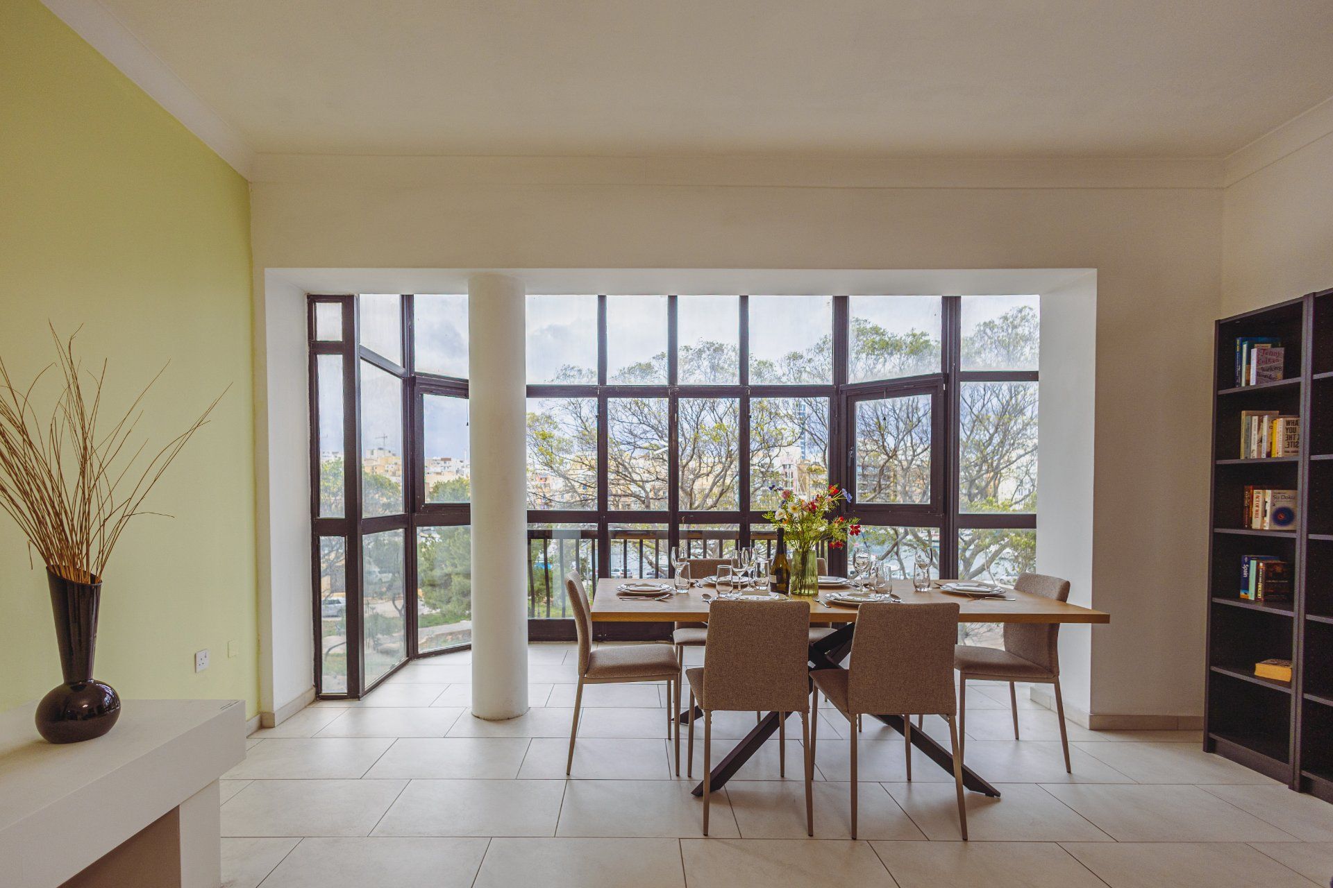 A dining area with a table set for four, large windows, and a bookshelf.