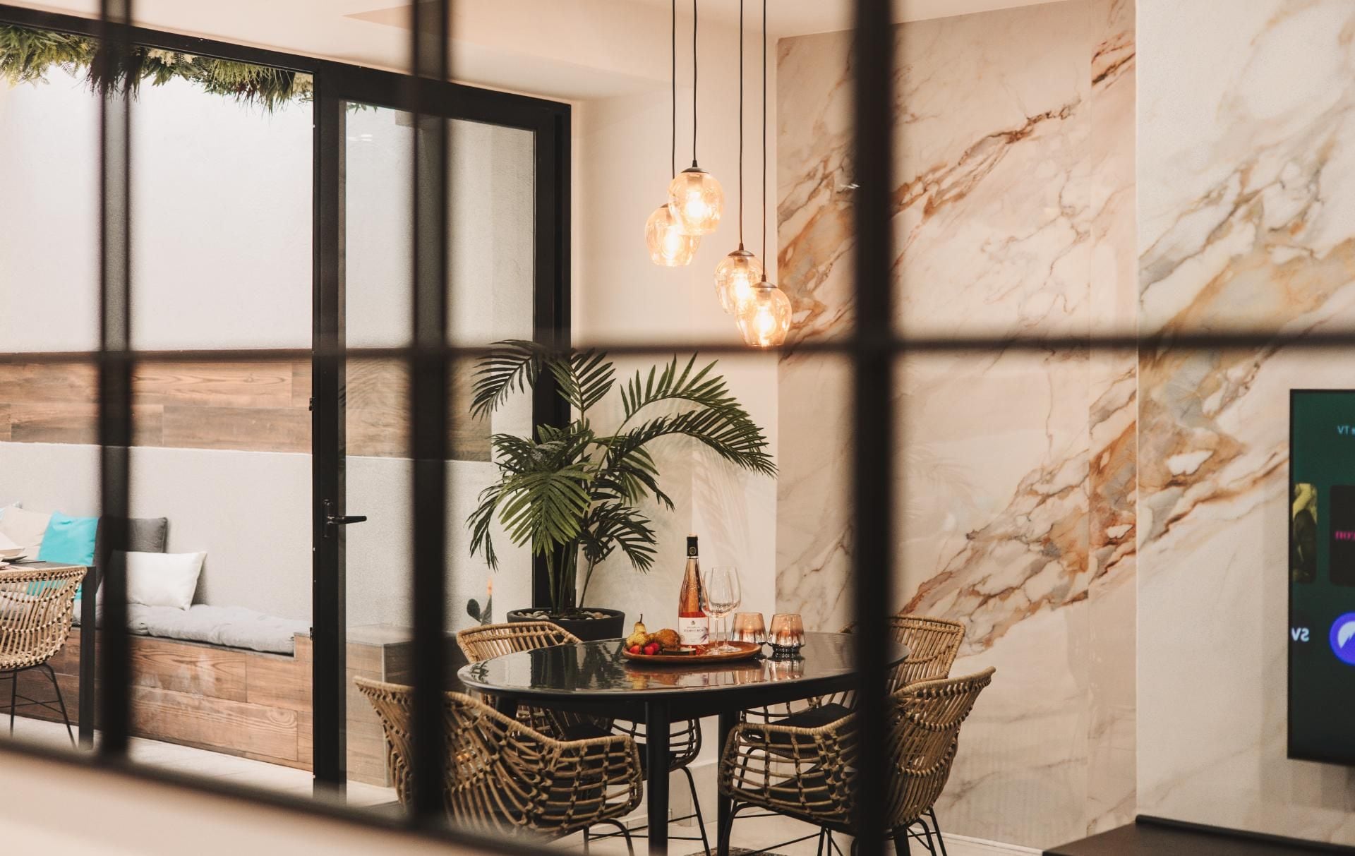 A dining area with a marble wall, a glass table, wicker chairs, and hanging pendant lights.