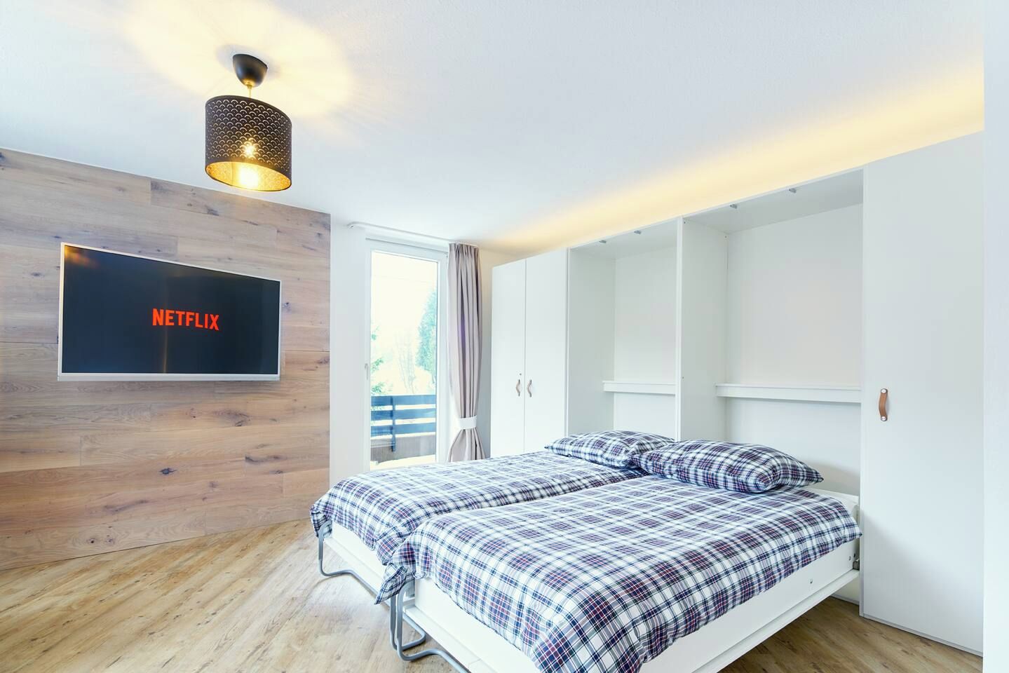 A modern bedroom with a bed, a wall-mounted TV displaying 'NETFLIX', and a wardrobe with sliding doors.