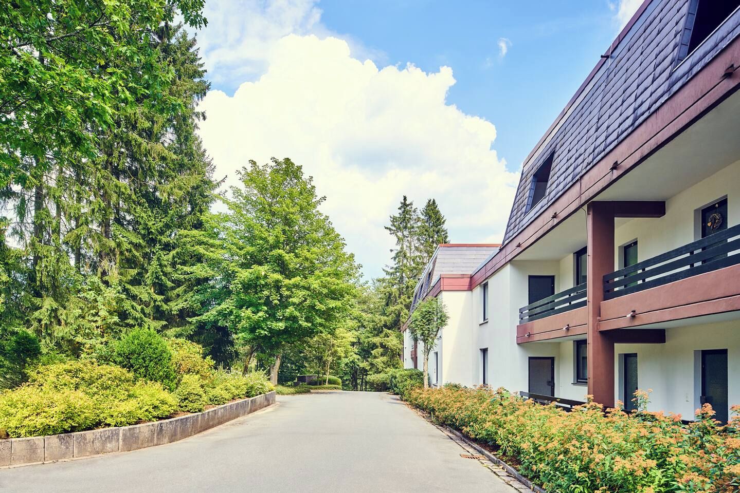 A building with multiple balconies, a paved driveway, and a row of trees.