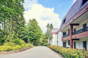 A building with multiple balconies, a paved driveway, and a row of trees. - Pool, sauna & ski jump view · #11 (Winterberg)