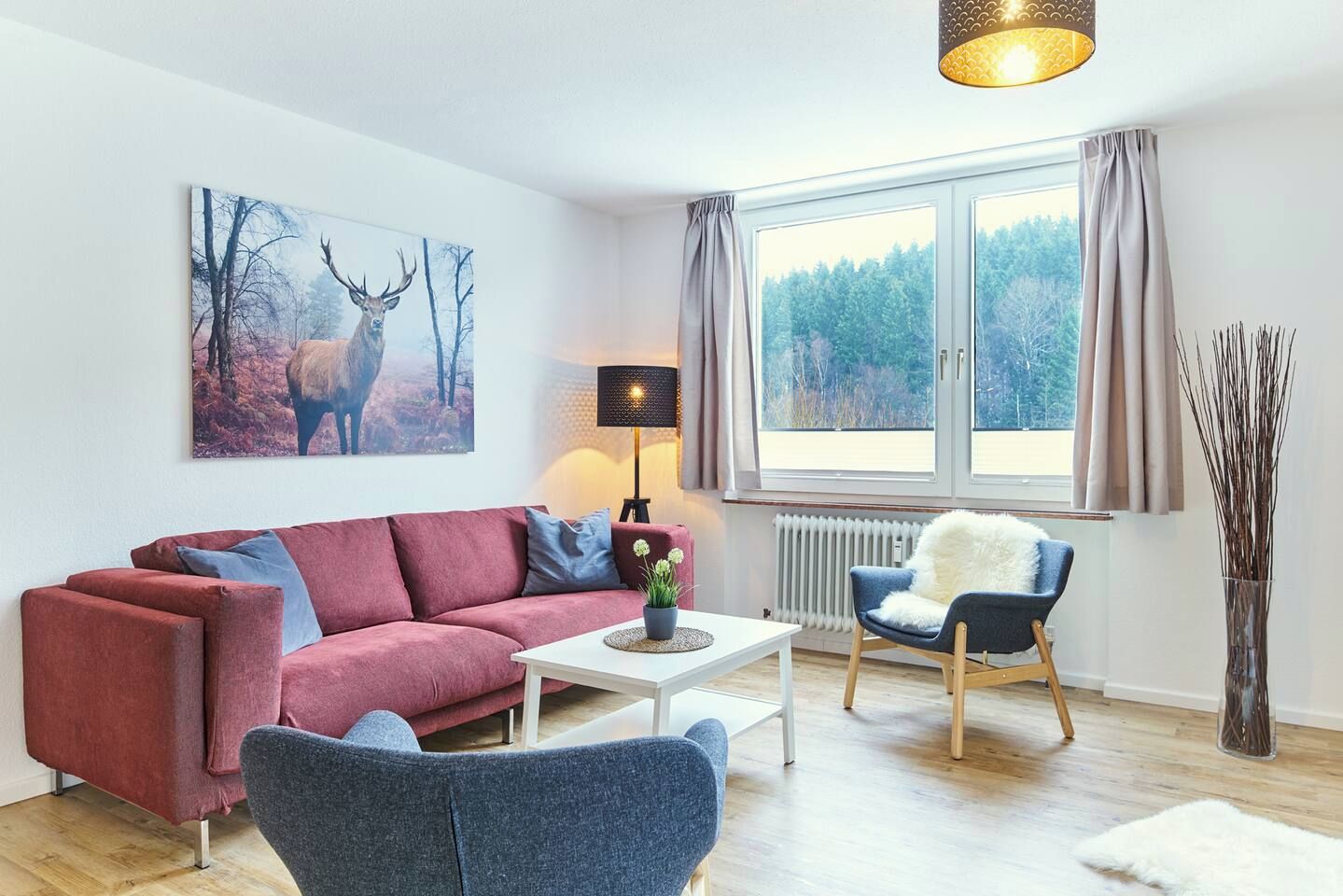 Sala moderna com sofá vermelho, poltrona azul, mesa de centro branca e pintura de veados na parede.