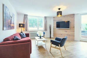 A modern living room with a red sofa, a blue armchair, a white coffee table, a floor lamp, a TV mounted on a wooden wall, and a large window with curtains. - Pool, sauna & ski jump view · #11 (Winterberg)