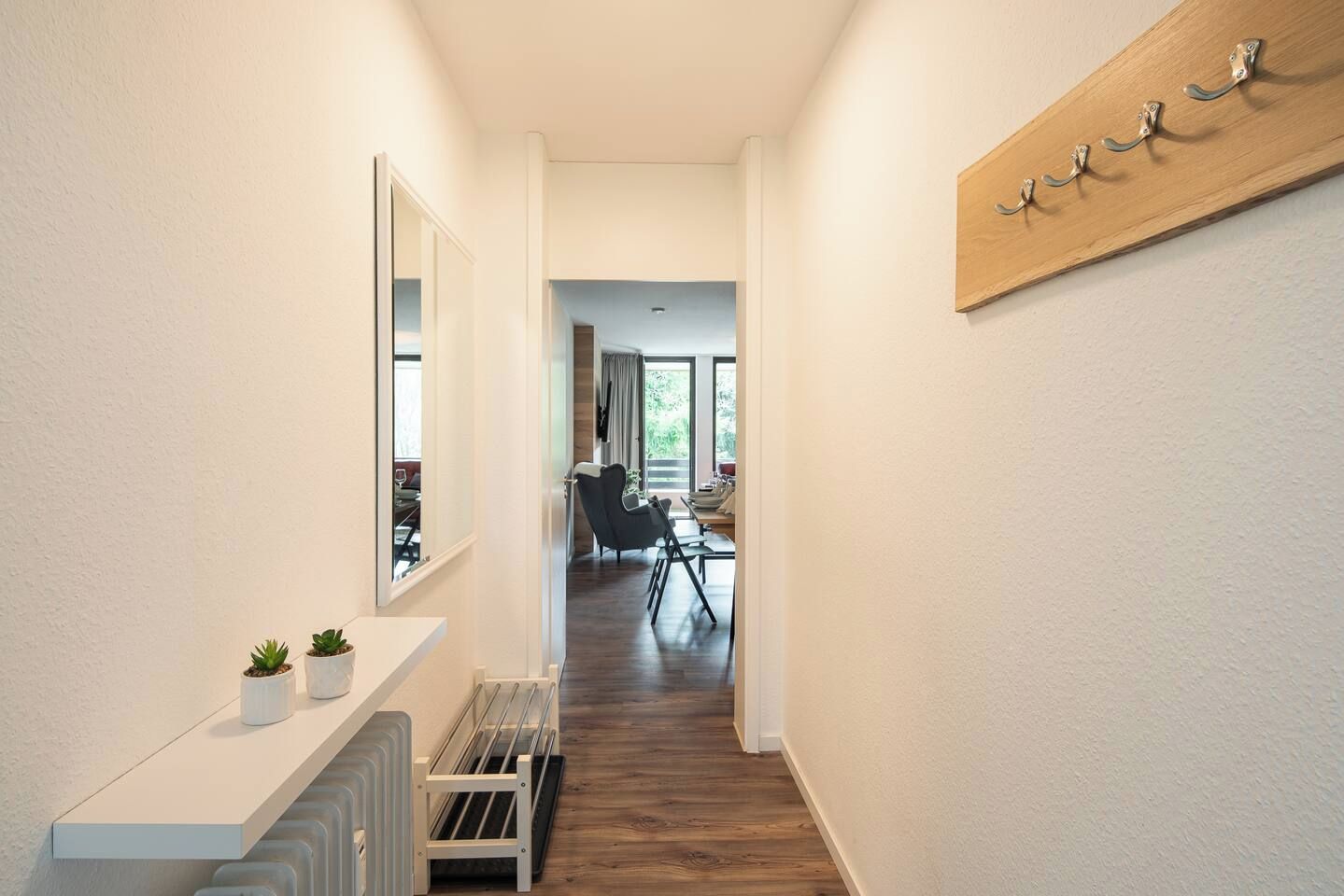 Ein Flur mit einem weißen Heizkörper, einer Bank und einem Spiegel links. Rechts befindet sich ein an der Wand befestigtes Holzgestell mit Haken.