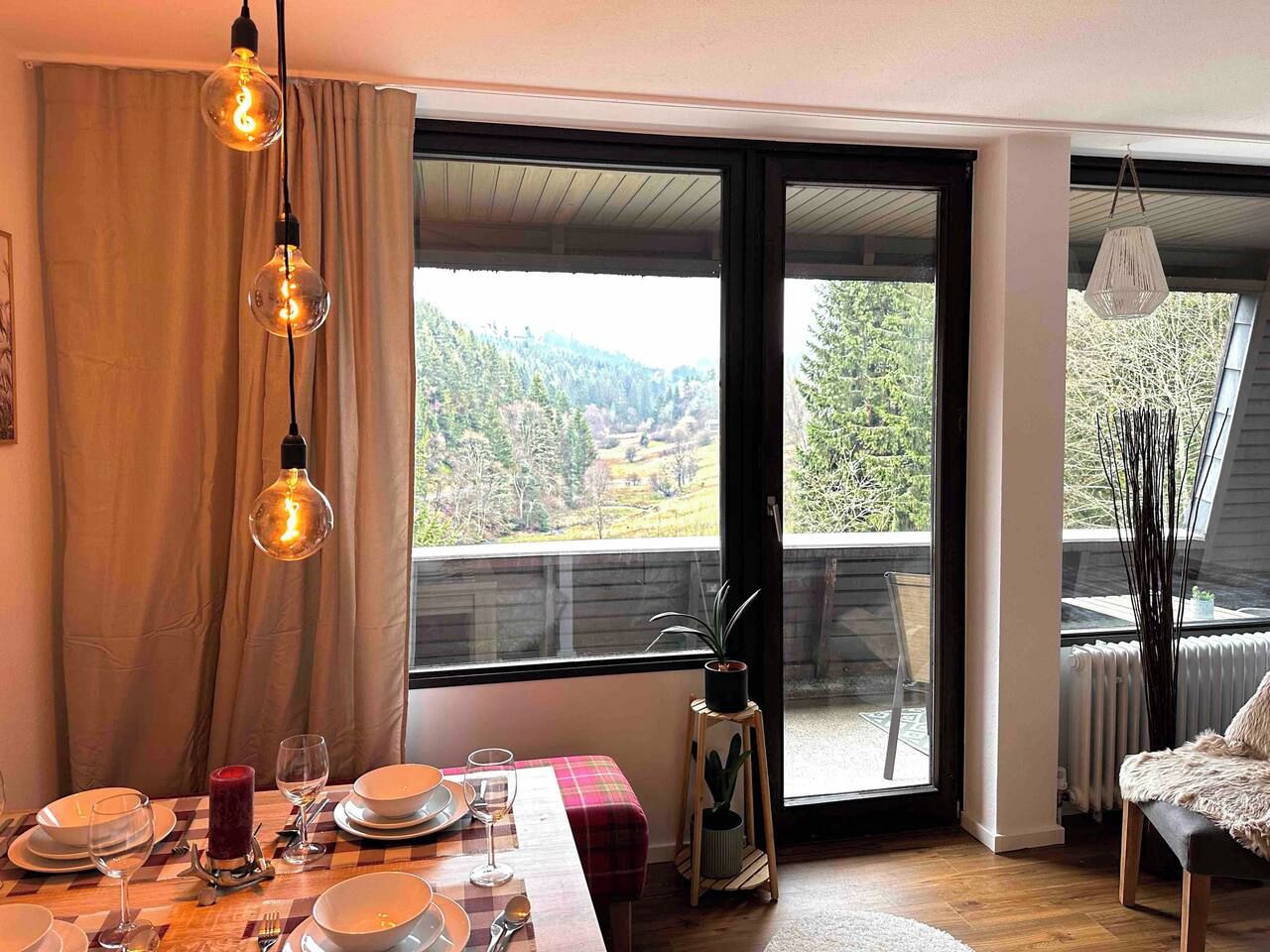 A dining area with a table set for four, a view of a forested landscape through a large window, and a balcony with a chair.