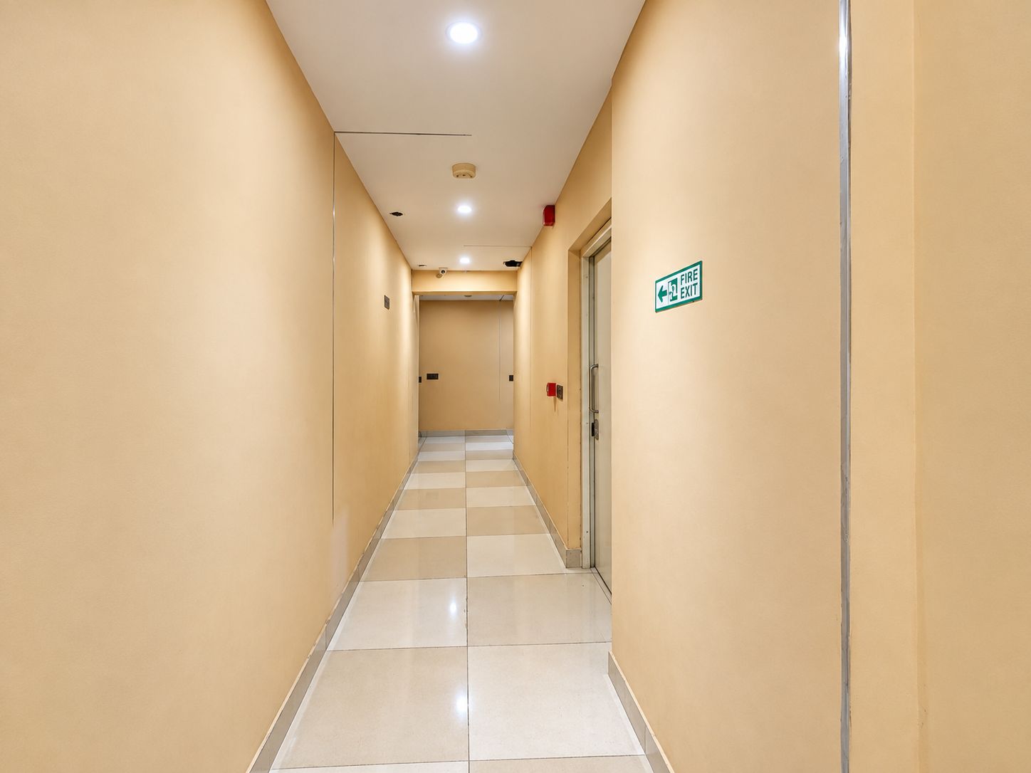 A hotel corridor with a checkered floor, beige walls, and a fire exit sign.