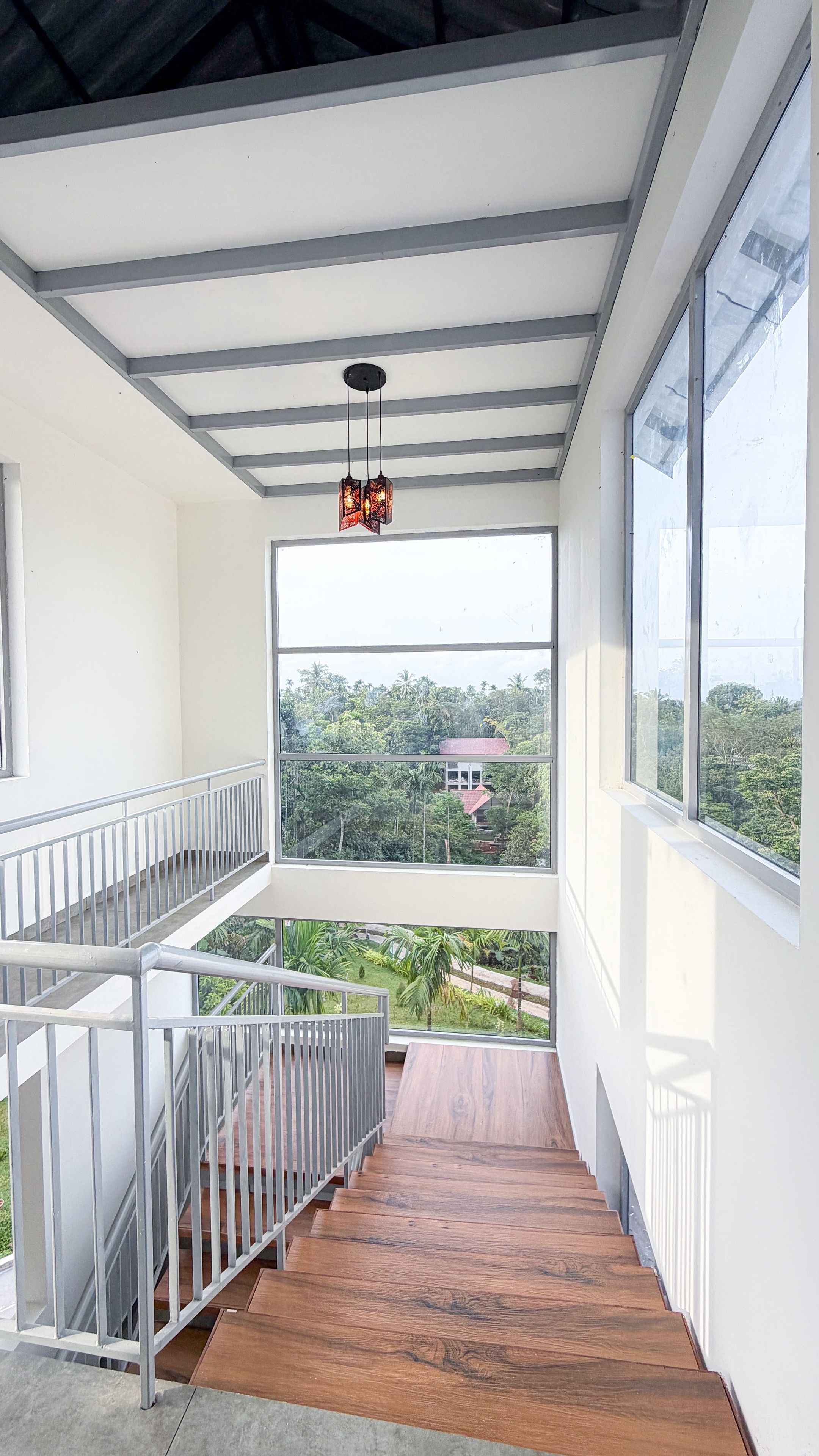 Un interior moderno con una escalera de madera, un balcón con barandilla metálica y un ventanal que ofrece vistas a vegetación y un edificio distante.