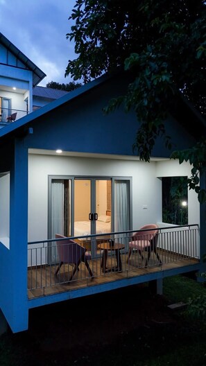 A modern two-story building with a balcony, a sliding glass door leading to a bedroom, and a small outdoor seating area with chairs.
