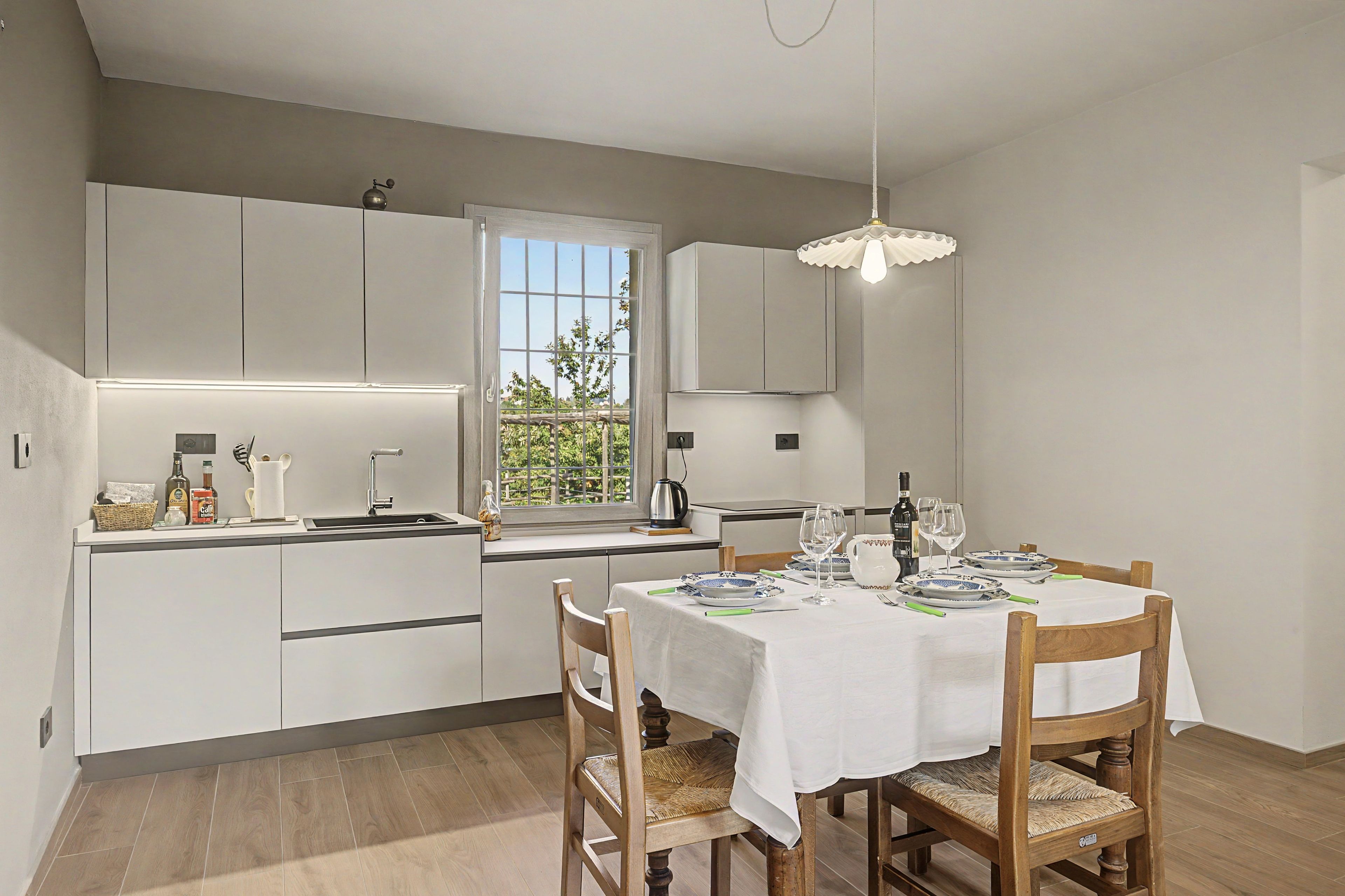 Une cuisine moderne avec des meubles blancs, une table à manger dressée pour deux personnes et une fenêtre donnant sur un paysage verdoyant.