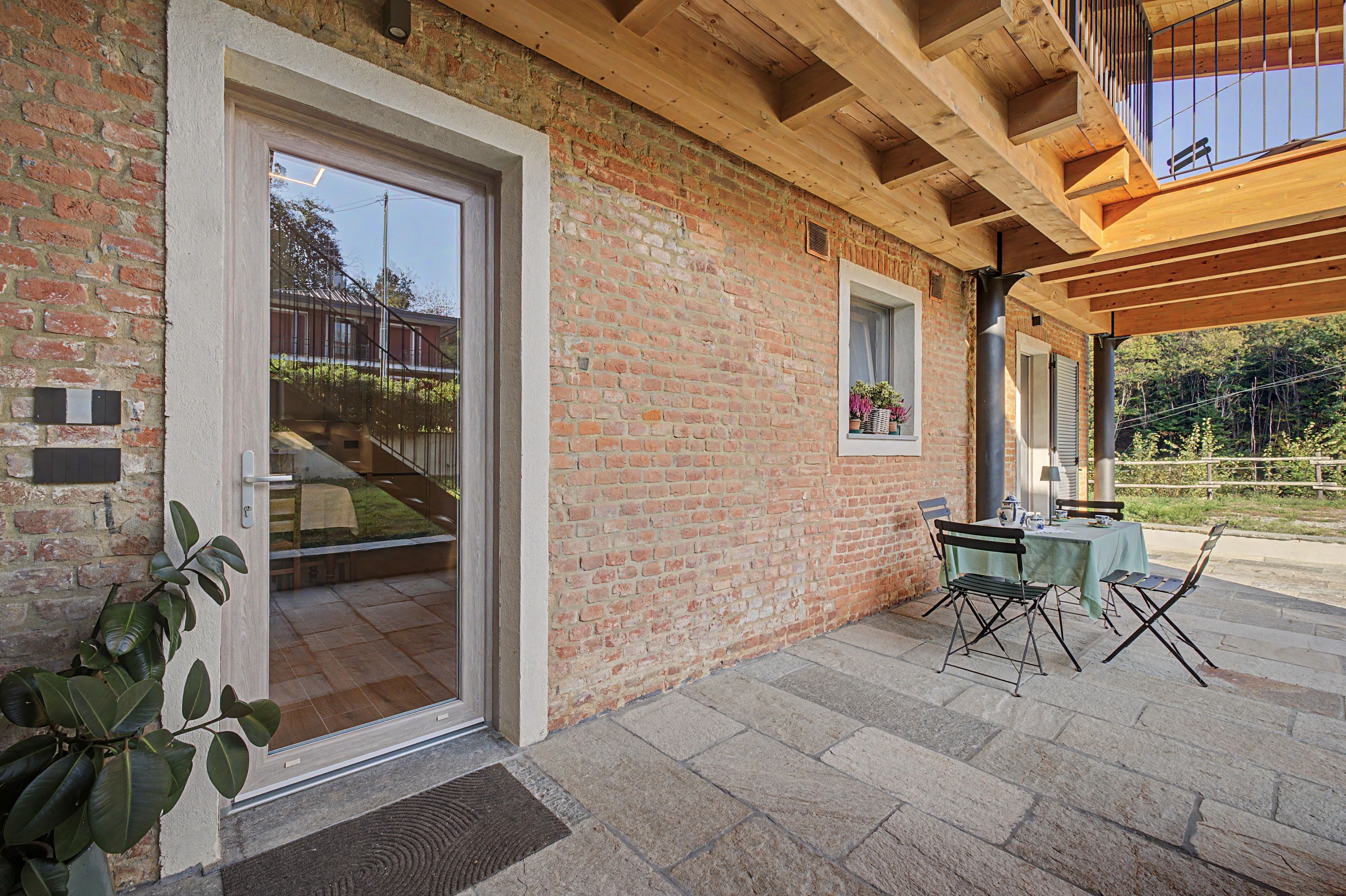 Une façade en briques avec une entrée dotée d’une porte en verre, une petite table avec des chaises à l’extérieur et une structure de plafond en bois.