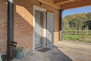 Un bâtiment en briques avec une porte fermée et une fenêtre fermée par des volets, un banc et un pot de fleurs posé sur le sol.