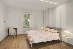 Una camera da letto con pavimento in legno, un letto con copriletto floreale, due abat-jour, una finestra con tende e un piccolo tavolino con una sedia.