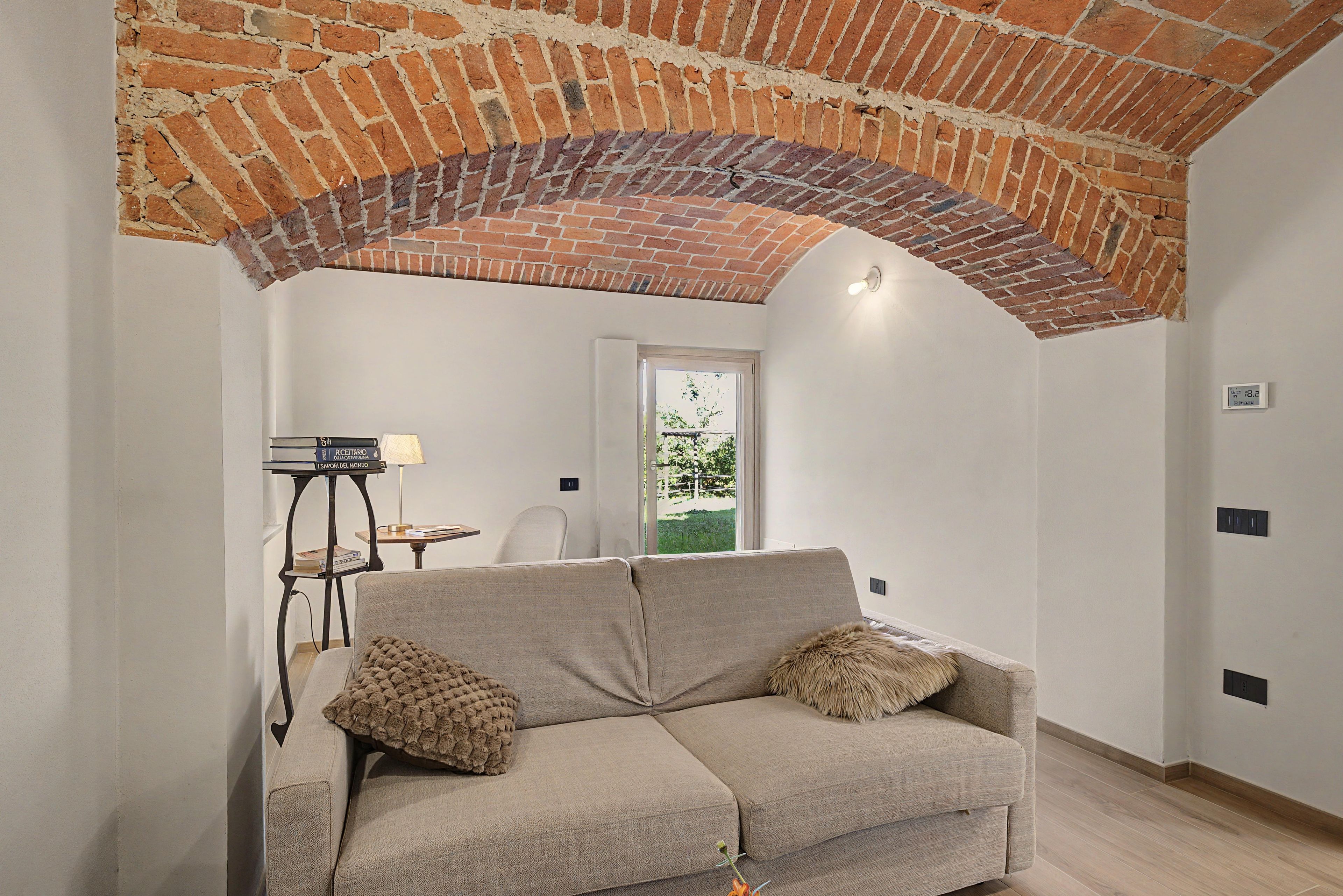 Un salon avec un plafond en briques, un canapé, une lampe et une vue sur l’extérieur, visible à travers une porte.