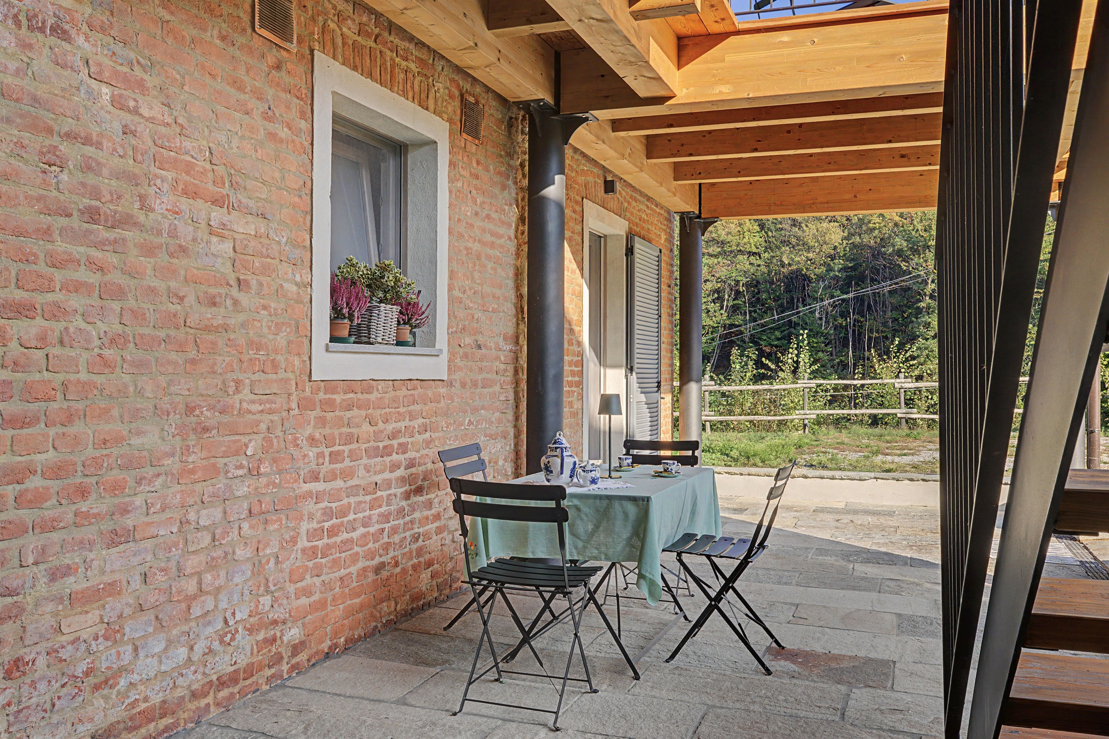 Un patio exterior con muro de ladrillo, una ventana con plantas, una mesa con sillas y una pérgola de madera.