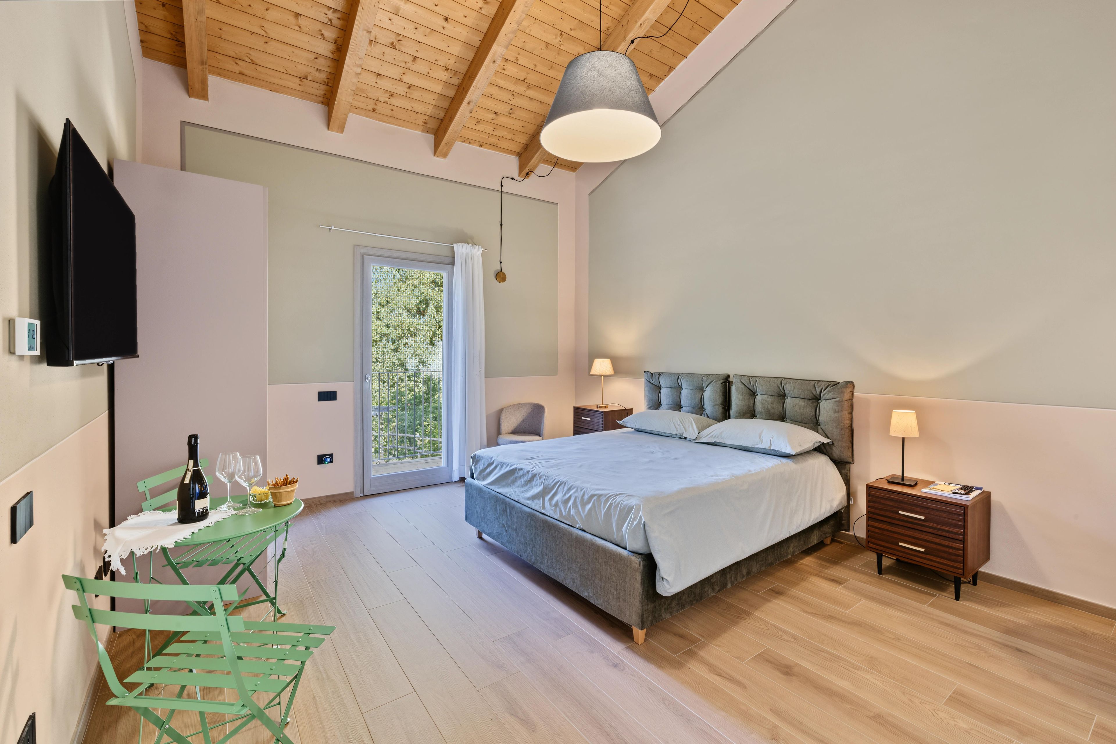 Une chambre à coucher avec un lit, des tables de chevet, un téléviseur fixé au mur, une fenêtre avec des rideaux et un fauteuil vert.