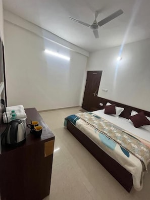 A hotel room with a bed, a desk with a kettle and cups, a ceiling fan, and a door. - Hotel dayalu palace (Vrindavan)