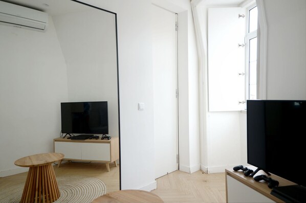 A modern, minimalistic room with a flat-screen TV on a wooden stand, a small round wooden table, and a large window providing natural light. - New Listing (Lisboa)