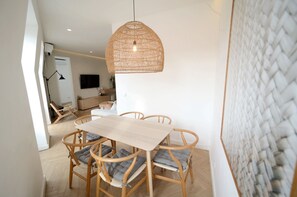 Un coin repas avec une table et des chaises en bois, un luminaire suspendu en fibres tressées et un téléviseur en arrière-plan.