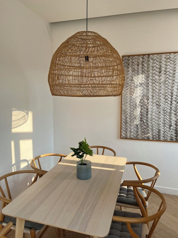 Un coin repas comprenant une table ronde en bois, quatre chaises, un luminaire suspendu en rotin et une œuvre d’art tissée accrochée au mur.