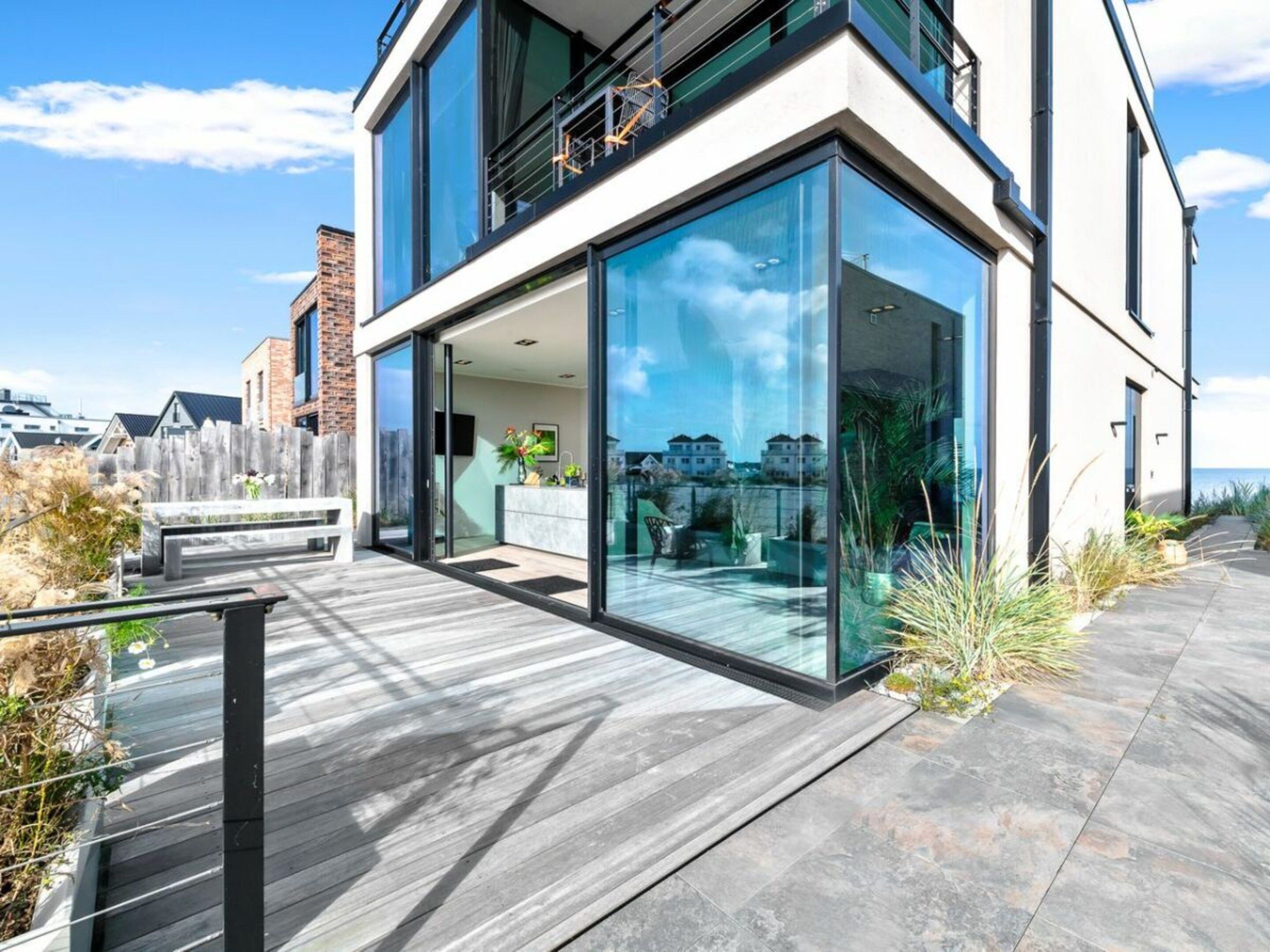 Un bâtiment moderne à deux étages, doté de grandes fenêtres en verre, d’une terrasse en bois et offrant une vue sur la mer.