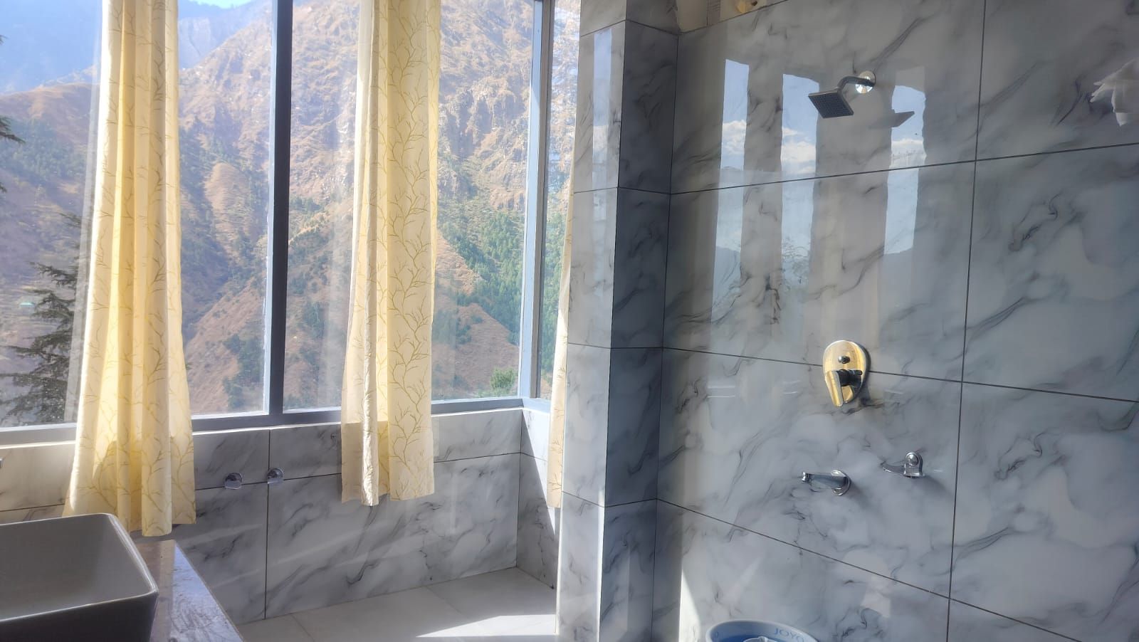 Een badkamer met een marmeren wand, een douche met een gouden kraan en een raam met gordijnen.