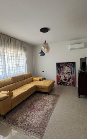 A living room with a sectional sofa, a patterned rug, a painting, and a television on a stand. - New Listing (Vittorio Veneto)