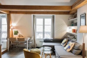 A cozy living room with a sofa, a small table, and a chair. There is a window with curtains, a bookshelf, and a lamp. -  Studio Apartment Overlooking Luxembourg Gardens - Latin Quarter (Paris)