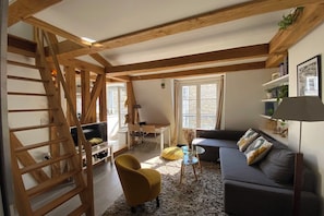 Un espacio habitable tipo loft con vigas de madera a la vista, un sofá, un sillón amarillo, una mesa de centro de vidrio y una escalera.