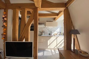 Habitación con techo de madera, televisión, lámpara y cocina visible a través de un umbral abierto.