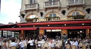 Um café animado ao ar livre, com mesas na calçada e um letreiro chamativo com a inscrição 'LE DEPART SAINT-MICHEL'.