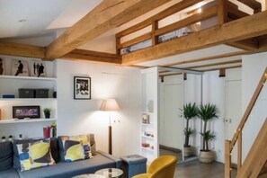 A modern living room with a loft area, wooden beams, a sofa, a coffee table, and a lamp. -  Studio Apartment Overlooking Luxembourg Gardens - Latin Quarter (Paris)