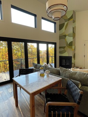 A modern living room with a large window, a fireplace, and a dining area. - The Room in the Woods. Hot Tub & Treetop Views. Collinsville, CT (Canton)