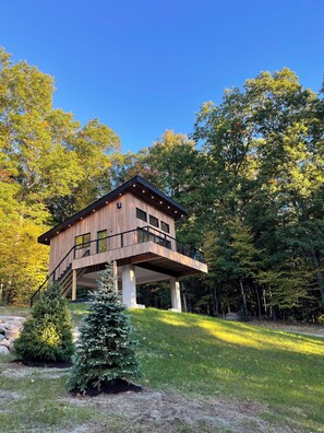 Une cabane en bois moderne dotée d’un balcon, entourée d’arbres et d’une zone herbeuse.