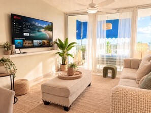 A living room with a flat-screen TV displaying a movie, a sofa, a coffee table, and a balcony with curtains. - COCO • Suite with Beach Access & Pool @ Isla Verde (Carolina)