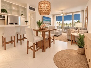 A modern living room with a dining area, a kitchen, and a large window with a view of palm trees. - COCO • Suite with Beach Access & Pool @ Isla Verde (Carolina)