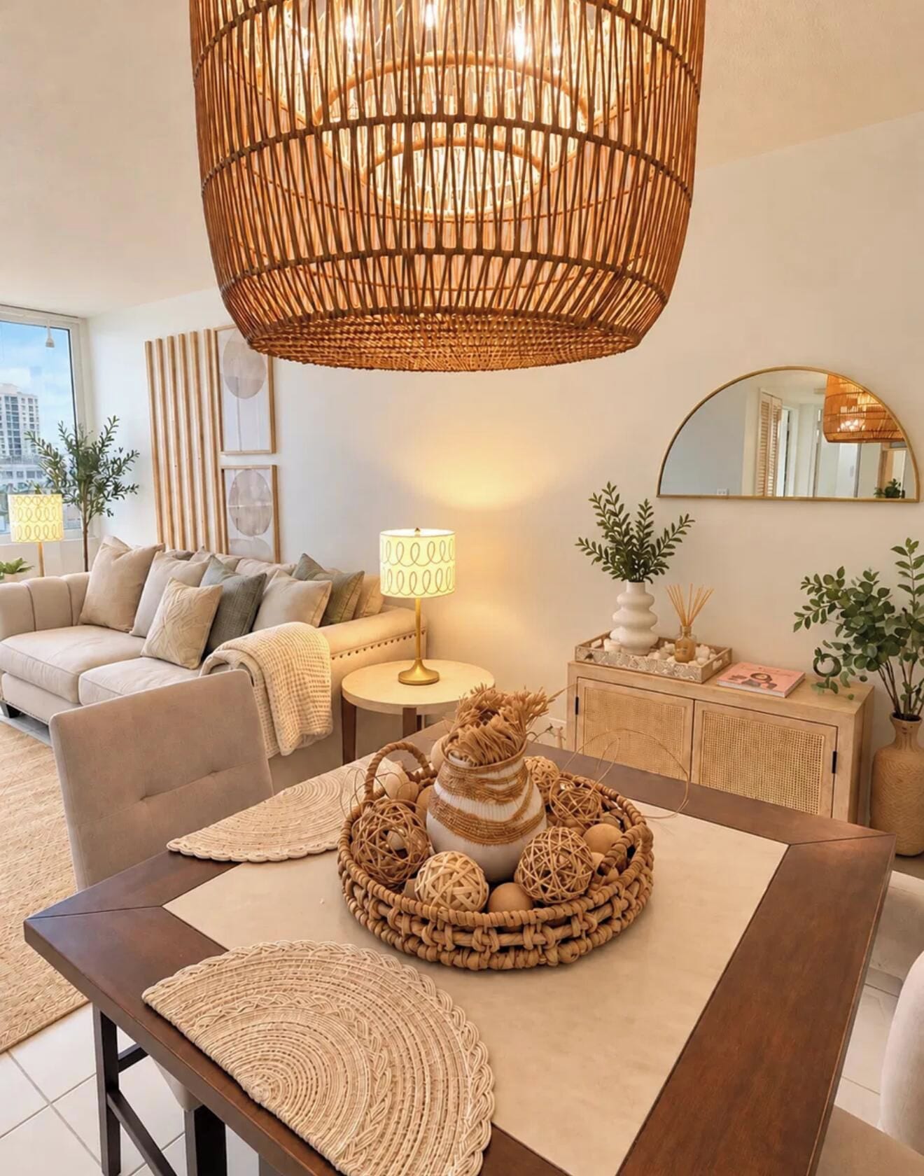 A modern living room with a large woven ceiling light, a round coffee table with woven decorations, a sofa, a side table with a lamp, and a mirror on the wall.