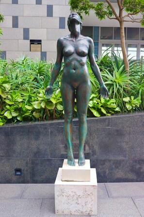 Une statue représentant une figure humaine, placée sur un piédestal, entourée de verdure et avec un bâtiment en arrière-plan.