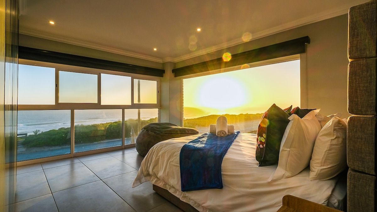 Une chambre avec un grand lit, une vue sur l’océan et un coucher de soleil.