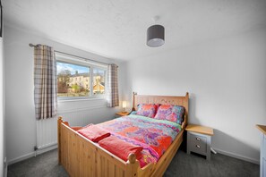 A bedroom with a wooden bed, a nightstand, a window with curtains, and a view of buildings outside. - Whole House In The Yorkshire Dales Near Harrogate (Dacre Banks)