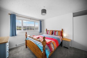 Ein Schlafzimmer mit einem hölzernen Bett, einem Fenster mit Vorhängen, einem Heizkörper, einem Nachttisch mit Lampe und einem Kleiderschrank.